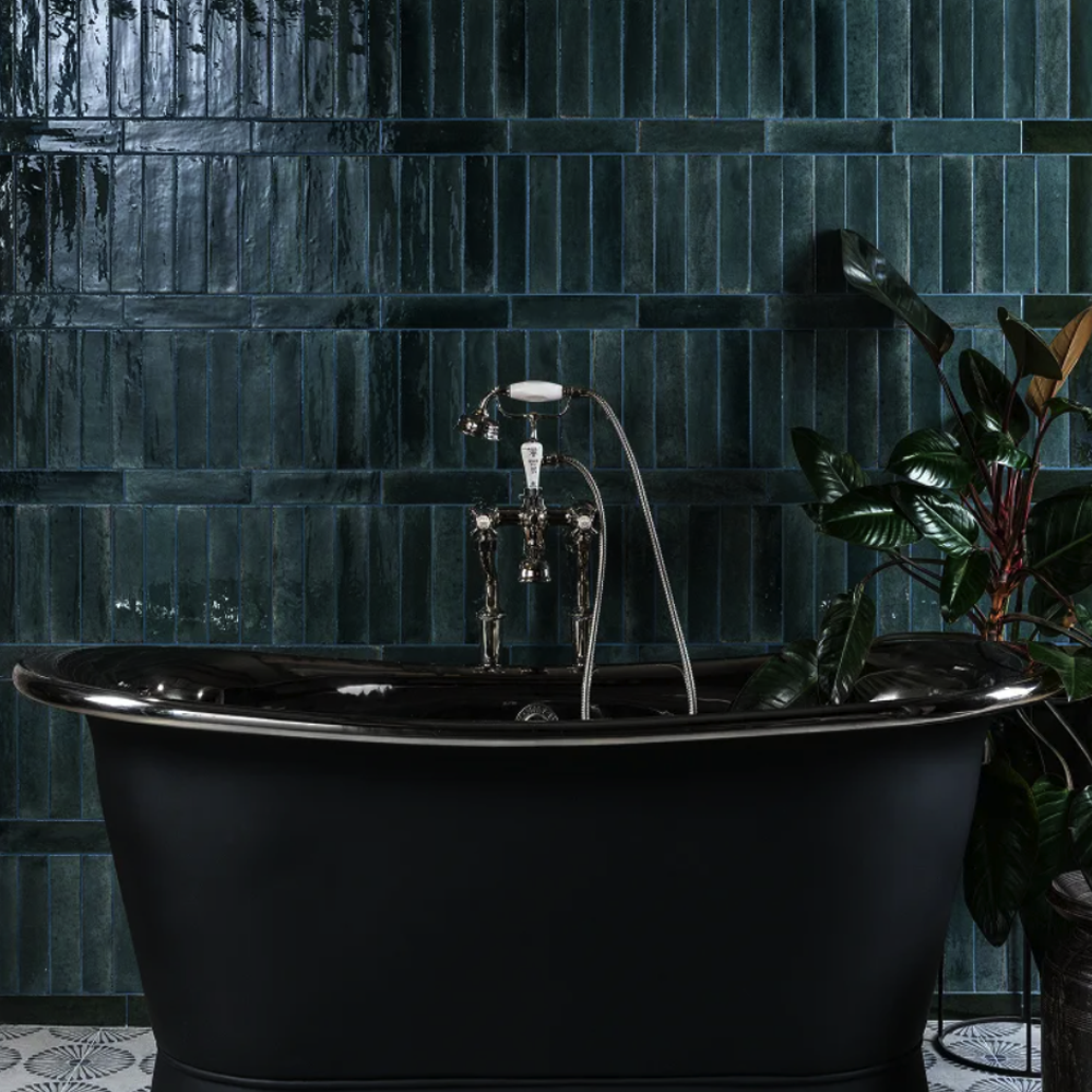 Black freestanding bathtub in a bathroom with dark tiled walls and a plant.