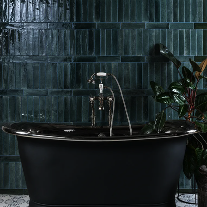 Black freestanding bathtub in a bathroom with dark tiled walls and a plant.